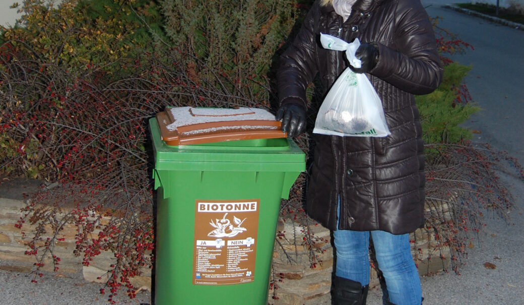 Winterlich gekleidete Frau wirft Bioabfälle in die angeschneite Biotonne.